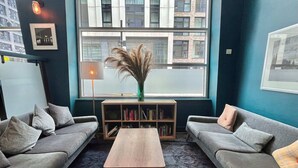A modern living room with two grey sofas, a wooden coffee table, a bookshelf, and a large window with a view of buildings. - Novotel Glasgow Centre (Glasgow)