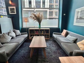 Una sala moderna con dos sofás grises, una mesa de centro de madera, una estantería y un ventanal con vistas a edificios.