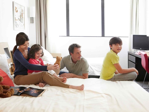 Una familia de cuatro personas sentada en una cama de un cuarto de hotel, con un escritorio y un televisor.