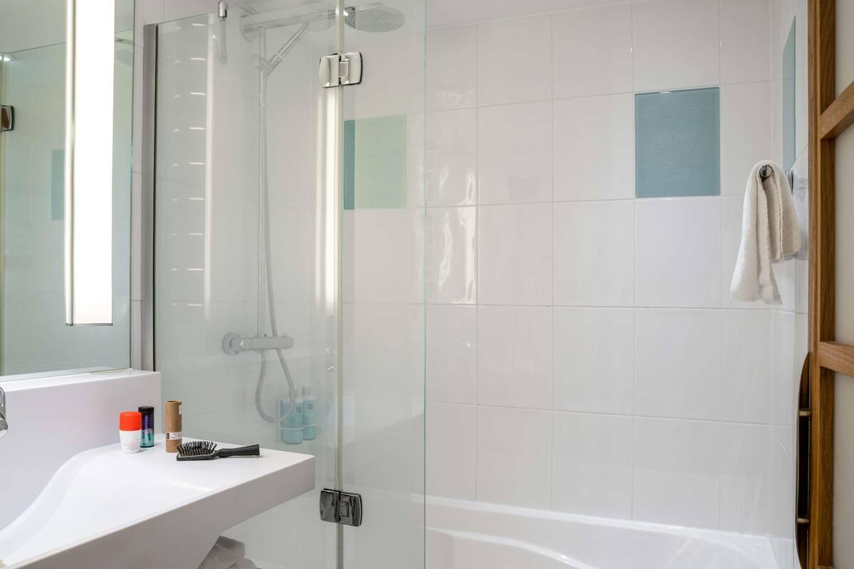 Un baño moderno con ducha envuelta en vidrio, lavamanos blanco con artículos de aseo y espejo con marco de madera.