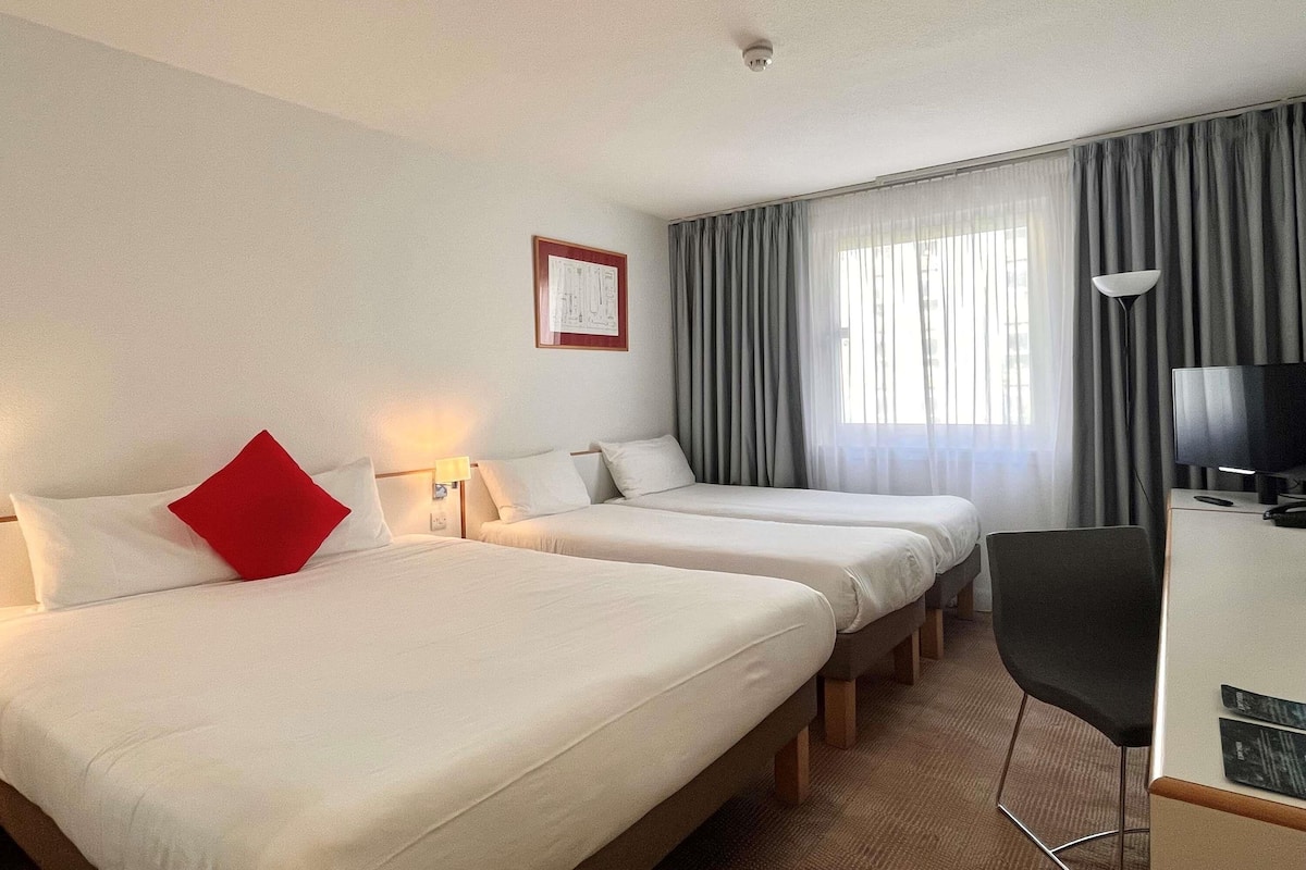 Ein Hotelzimmer mit zwei Betten, einem Schreibtisch mit Fernseher, einem Sessel und einem Fenster mit Vorhängen.