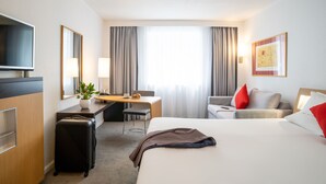 A hotel room with a bed, a desk, a chair, a TV, a lamp, and a suitcase. - Novotel Glasgow Centre (Glasgow)