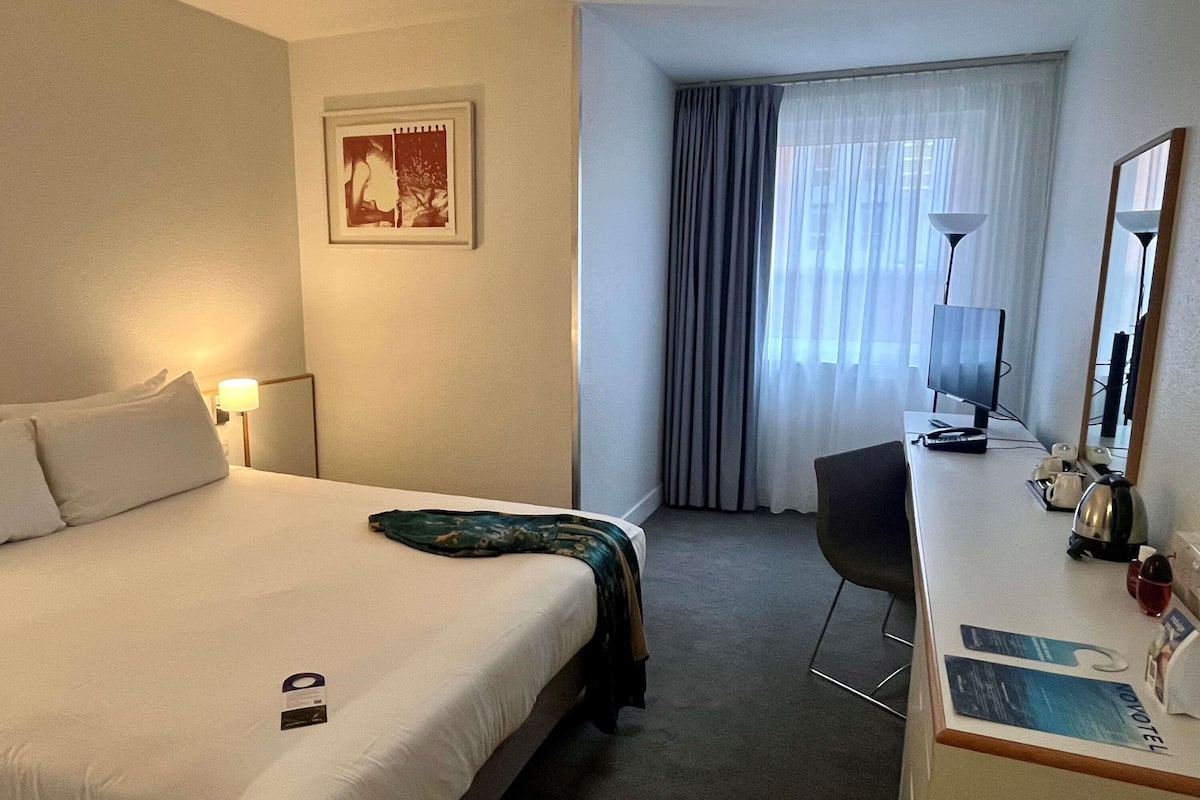 Ein Hotelzimmer mit Bett, Schreibtisch mit Computer, Lampe, Stuhl und einem Fenster mit Vorhängen.