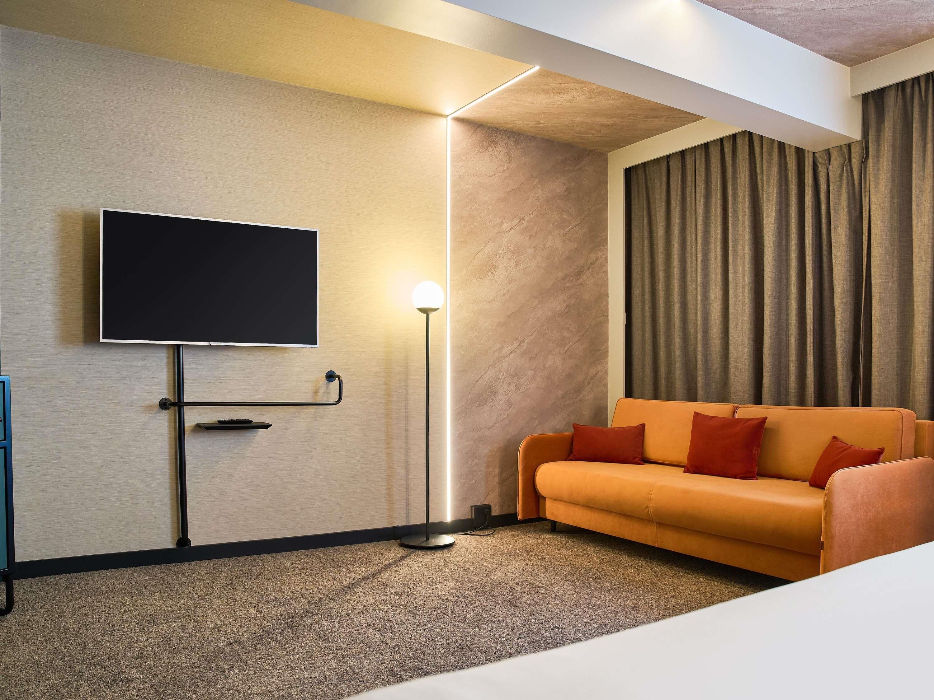 A hotel room with a flat-screen TV mounted on the wall, a floor lamp, an orange sofa with red cushions, and a large window with curtains.