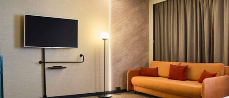 A hotel room with a flat-screen TV mounted on the wall, a floor lamp, an orange sofa with red cushions, and a large window with curtains.