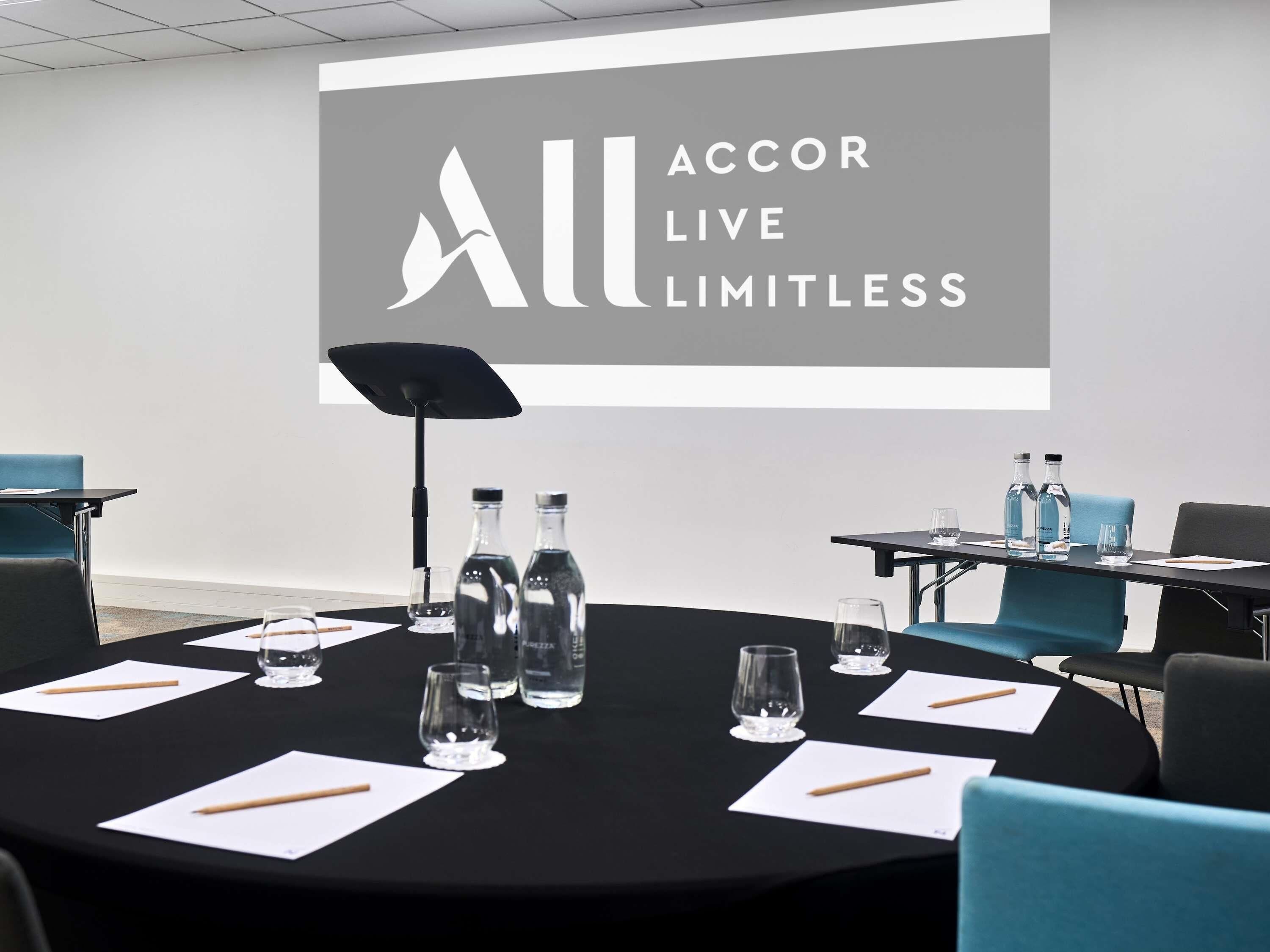 A conference room with a projector screen displaying 'ACCOR LIVE LIMITLESS', tables with water bottles, glasses, and notepads.