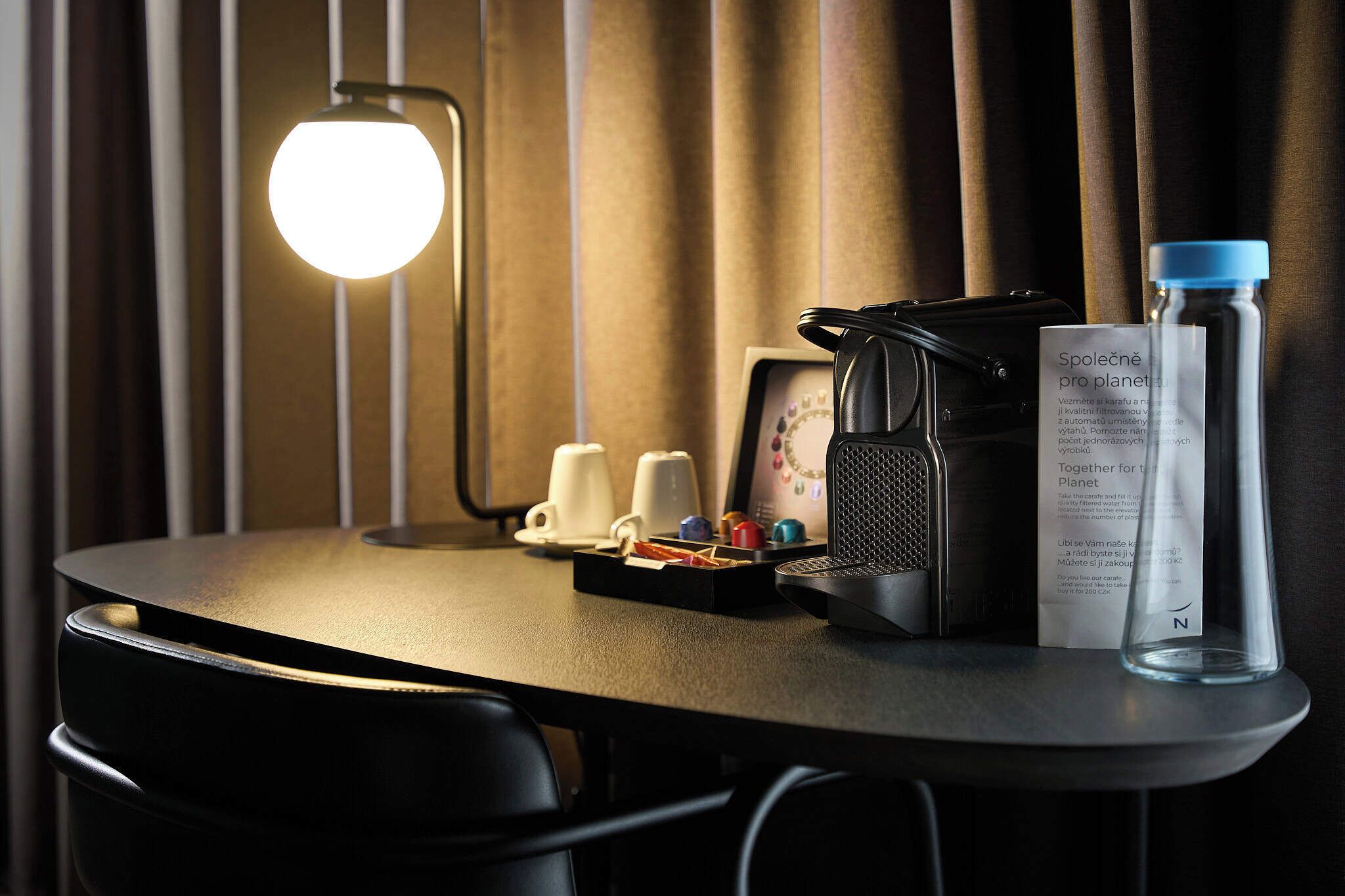 Et pænt hotelværelse med et rundt bord, en lampe, en bærbar computer, kaffemaskine og en vandflaske.
