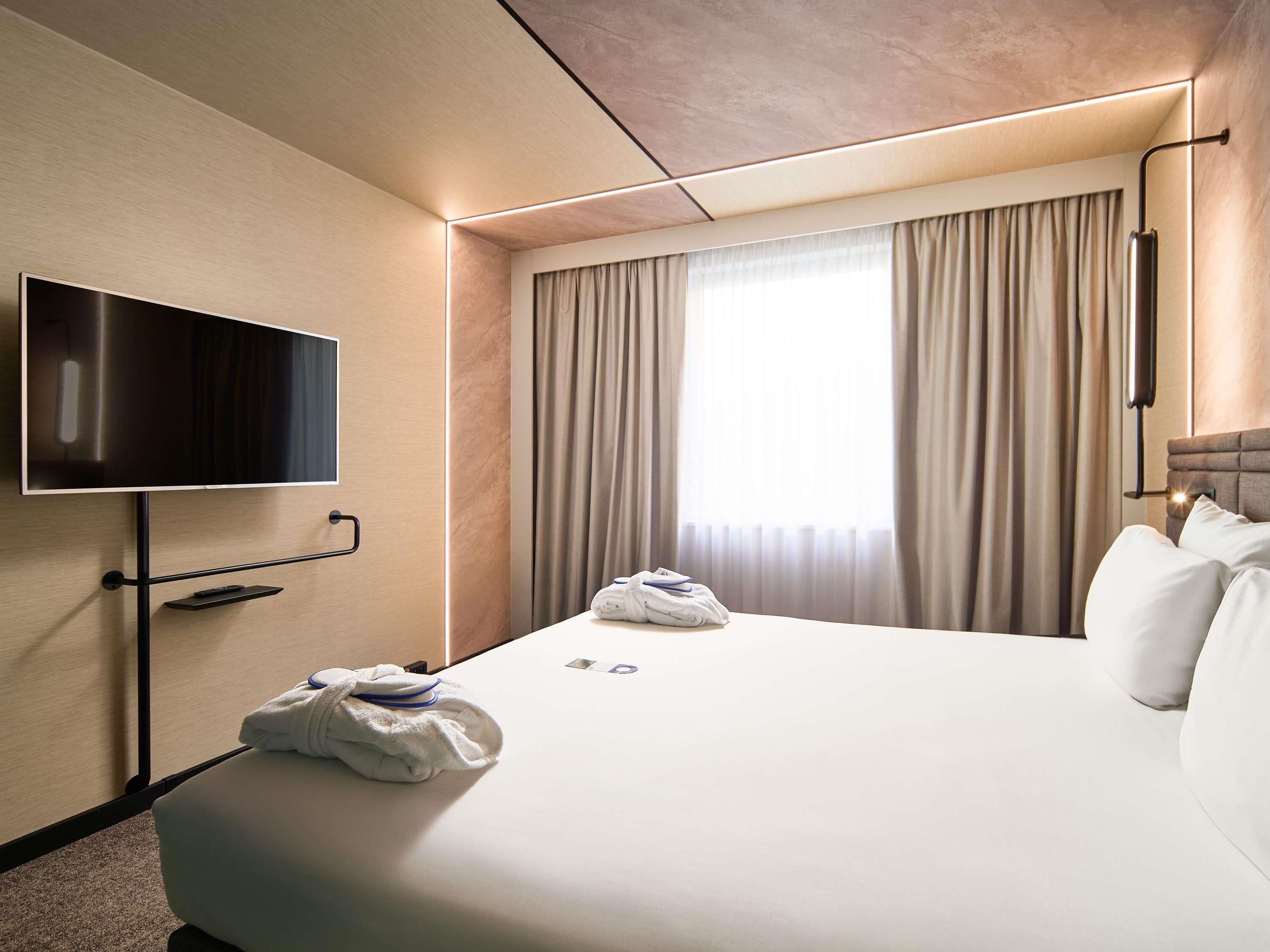 Una camera d'albergo con schermo piatto montato a parete, un letto con asciugamani ordinati, una finestra con tende e un comodino.