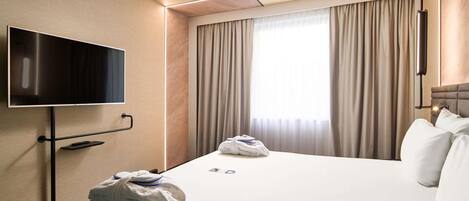 Habitación de hotel con televisor de pantalla plana en la pared, una cama con toallas ordenadas, una ventana con cortinas y una mesita de noche.