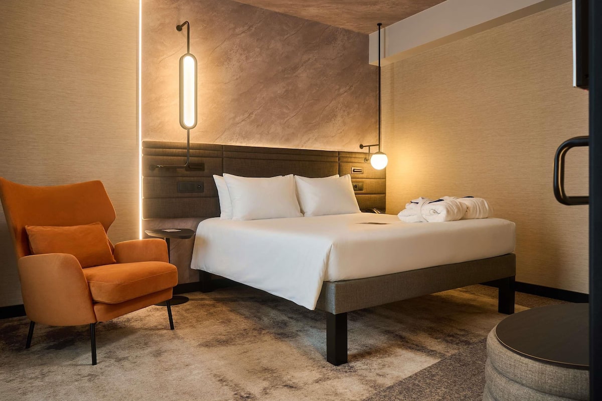 A modern hotel room with a bed, a chair, a bedside table, and a lamp.