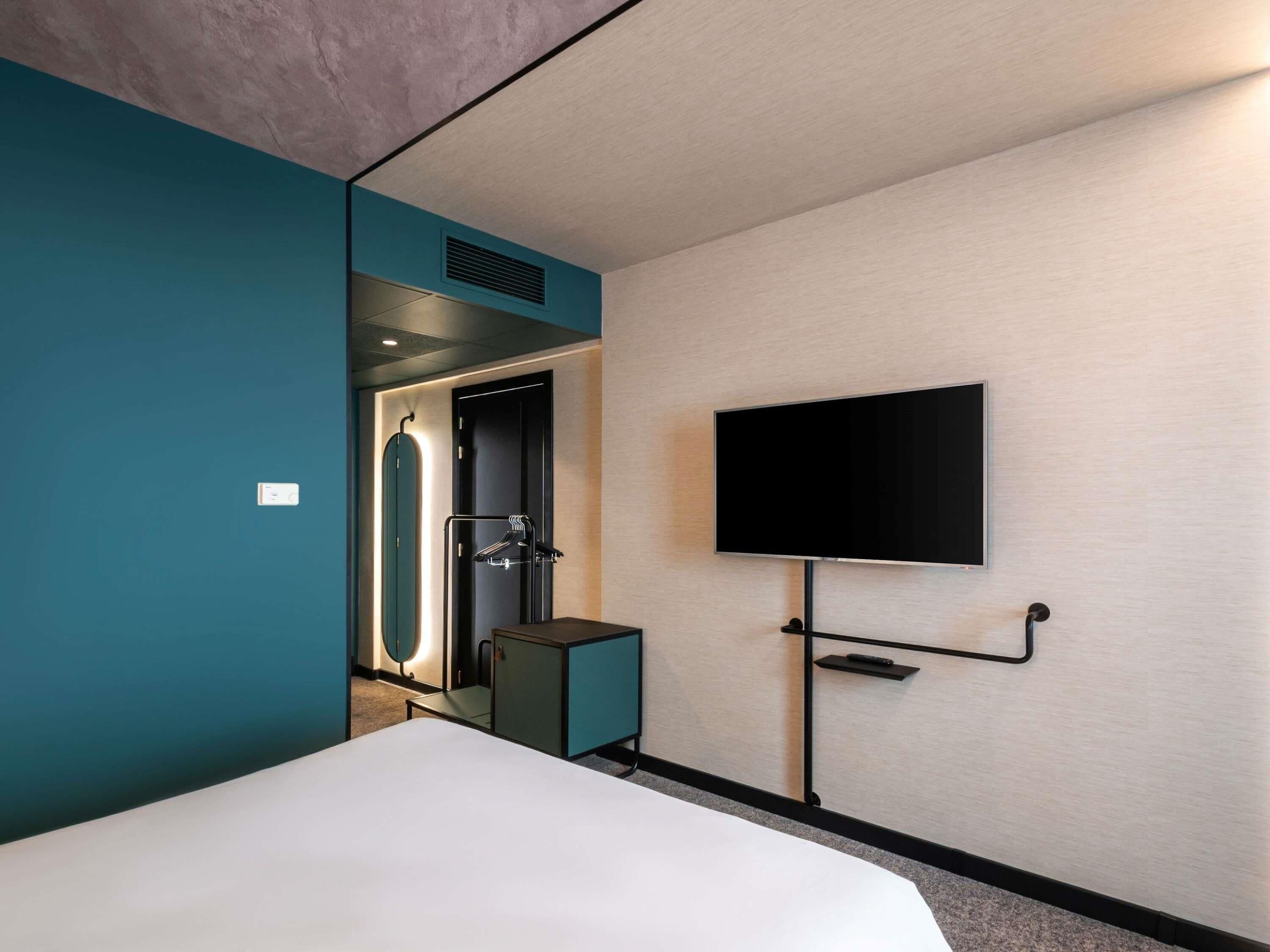 Quarto de hotel com uma cama, uma TV fixada na parede, uma porta com suporte para toalhas e um pequeno armário.