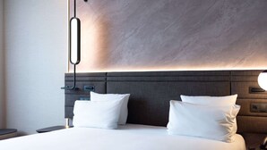Quarto de hotel moderno com uma cama grande, duas almofadas, uma mesinha de cabeceira e um candeeiro fixado na parede.