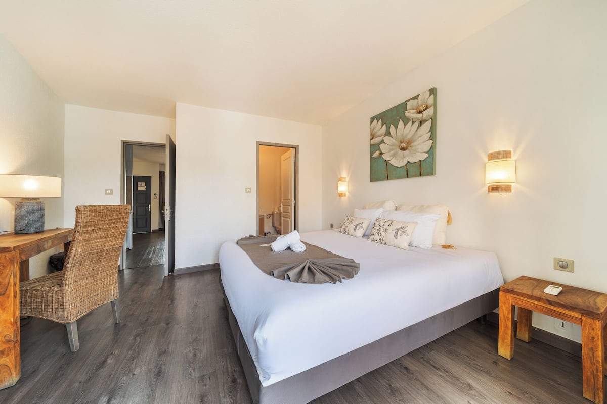 A hotel room with a large bed, a wooden chair, a small table, and a painting on the wall.