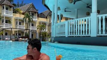 A man relaxing in a pool with a drink, surrounded by a resort building with balconies and a poolside area.