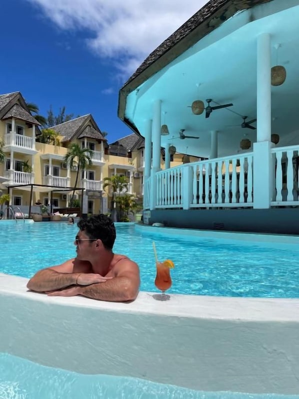 A man relaxing in a pool with a drink, surrounded by a resort building with balconies and a poolside area. - Hôtel Exsel Ermitage (Saint-Paul)