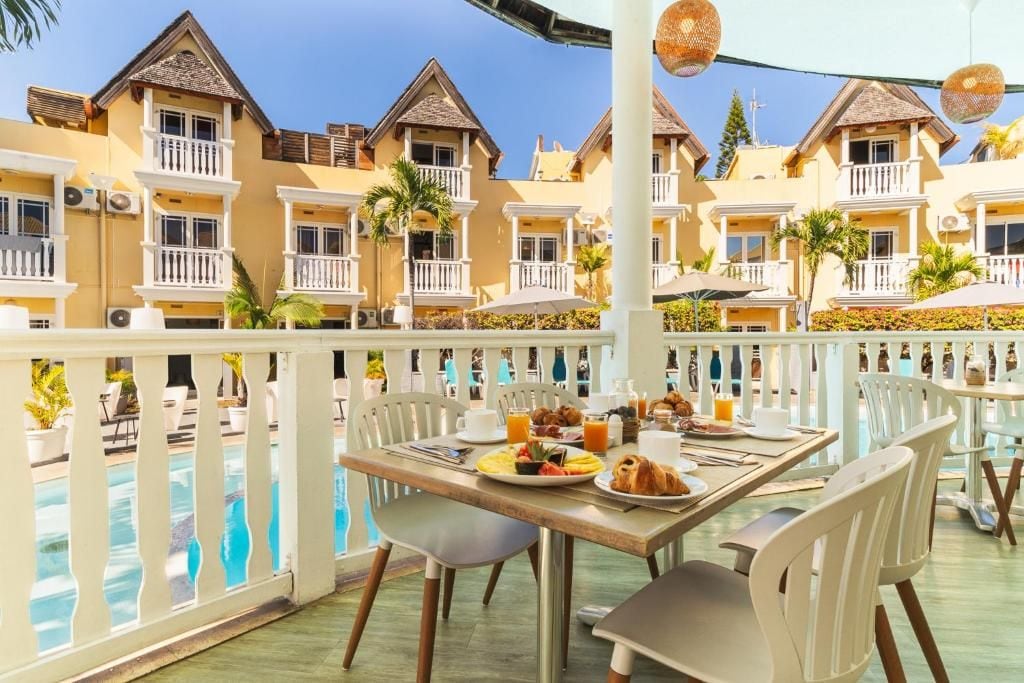 A hotel balcony with a pool, dining table with breakfast, and multiple-story buildings with balconies.