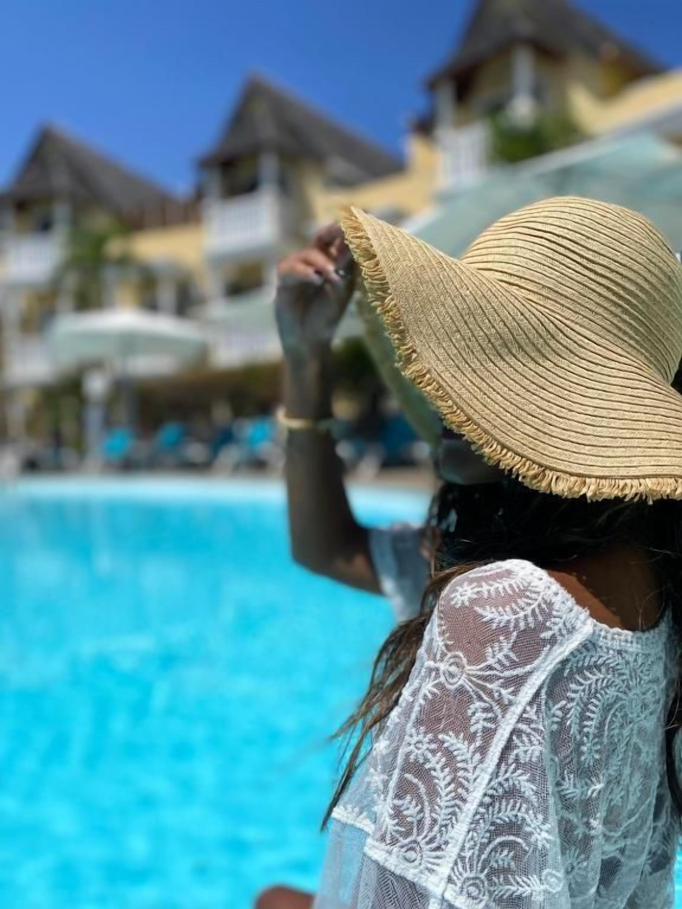 A person wearing a straw hat and a lace garment by a pool with thatched-roof buildings in the background.