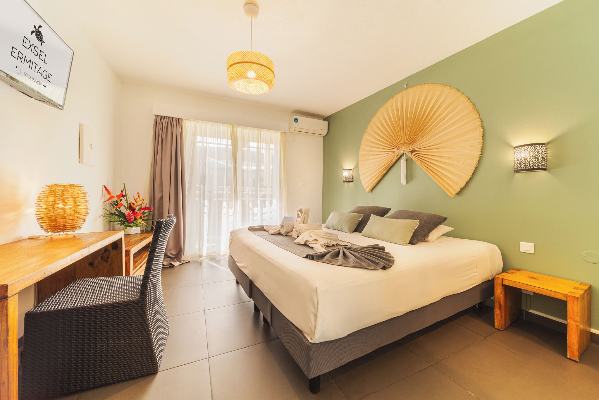 A hotel room with a bed, a desk, a chair, a fan, and a wall-mounted light fixture.