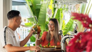 A person holding a drink at a bar with a plant in the foreground. - Hôtel Exsel Ermitage (Saint-Paul)