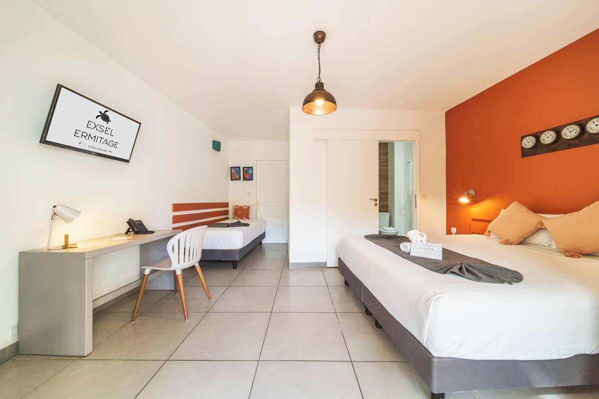 A hotel room with a desk, two beds, a chair, a desk lamp, a clock, and a framed sign on the wall.
