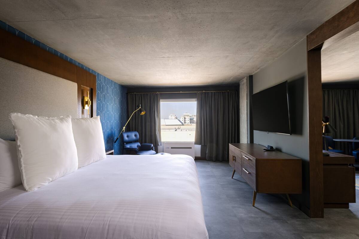 Habitación de hotel con una cama grande, un escritorio, un televisor de pantalla plana, una silla y una ventana con cortinas.