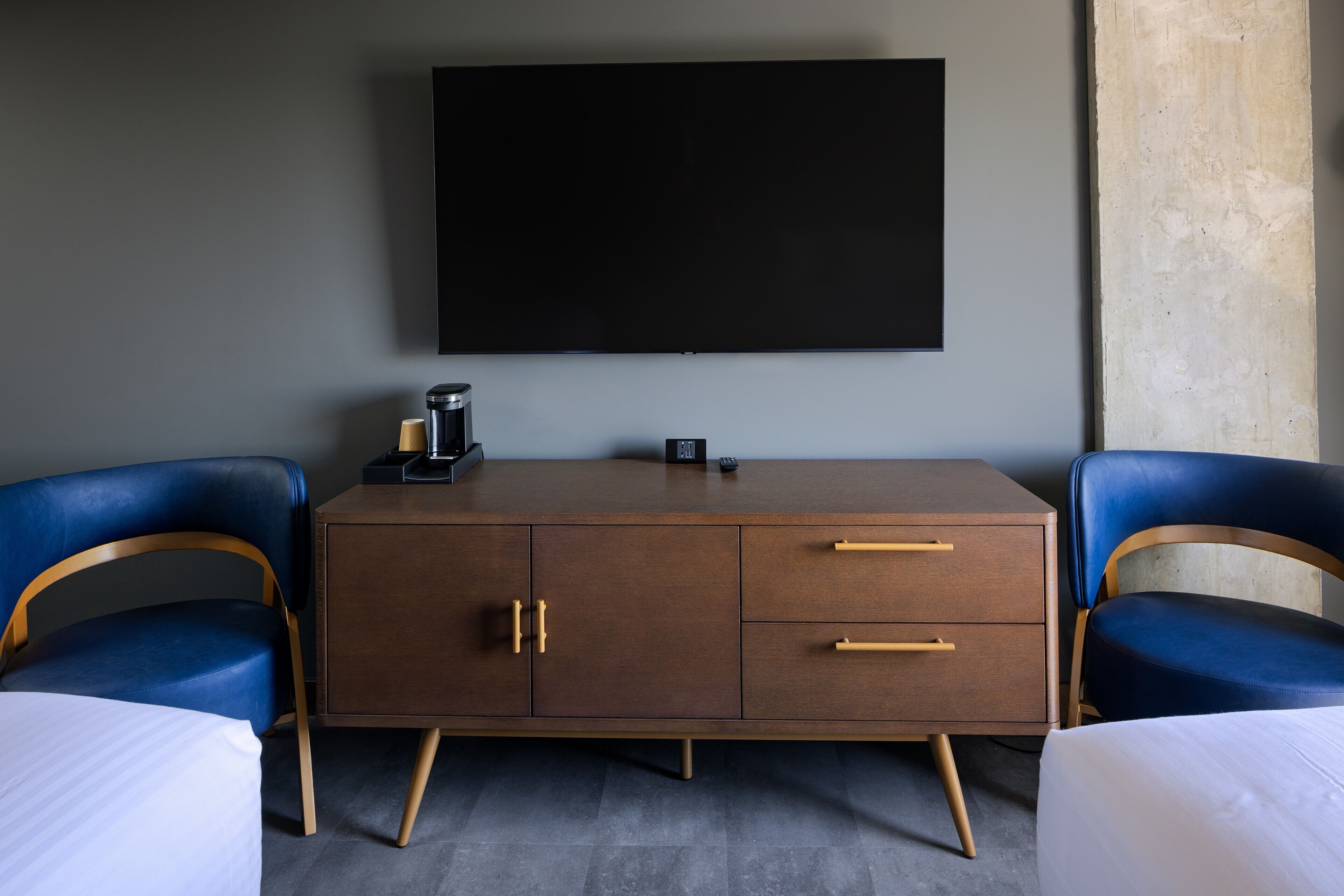 Et hotelværelse med et stort fladskærms-tv på væggen, et træskab med skuffer, to blå polstrede lænestole og en seng med hvid sengetøj.
