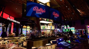Bar de casino com várias máquinas de jogo, área de bar com garrafas e placas indicando Bally's Baton Rouge Casino & Hotel.