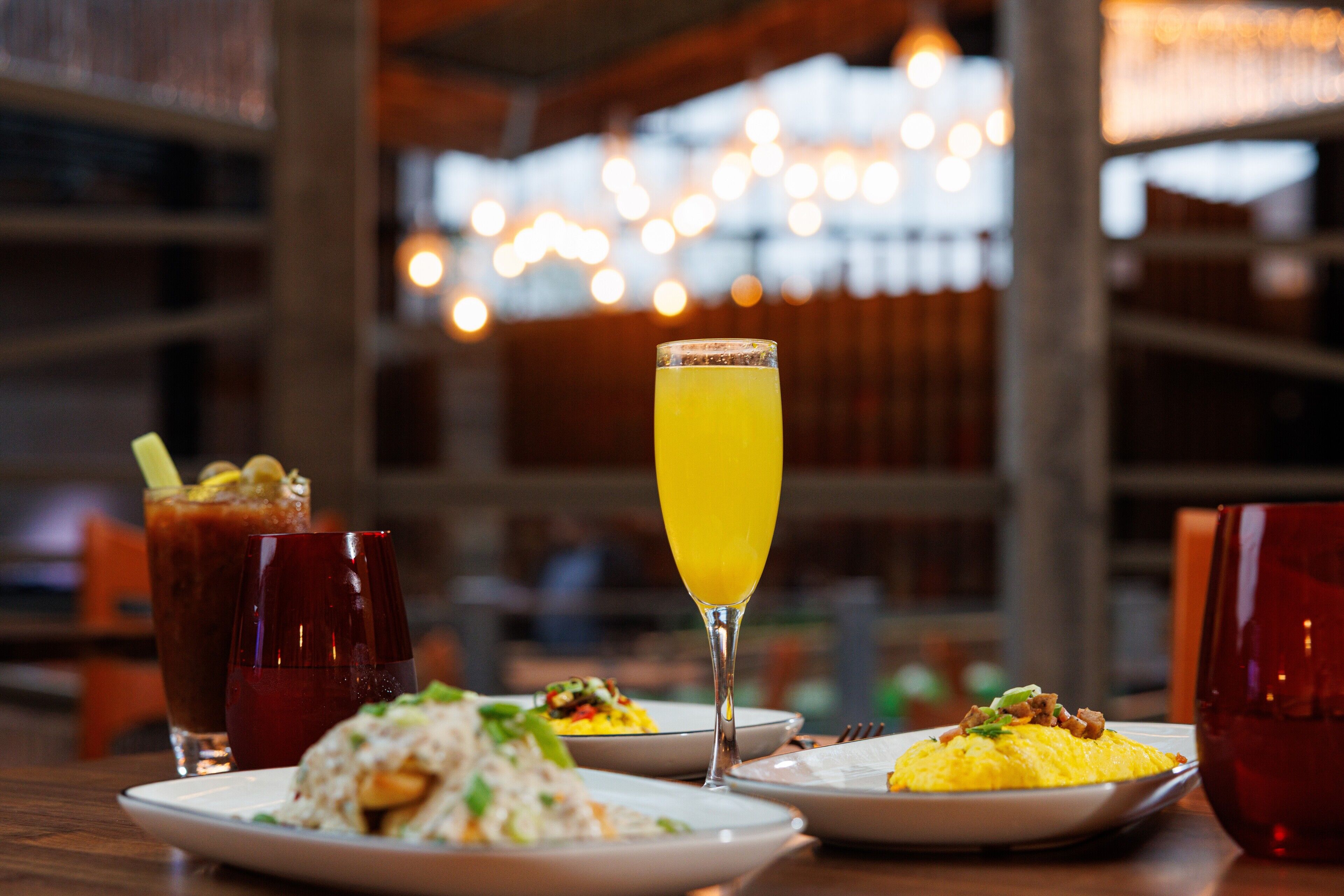 A dining table with various dishes and drinks, including a glass of orange juice and a red drink, in a restaurant setting with a blurred background of lights.