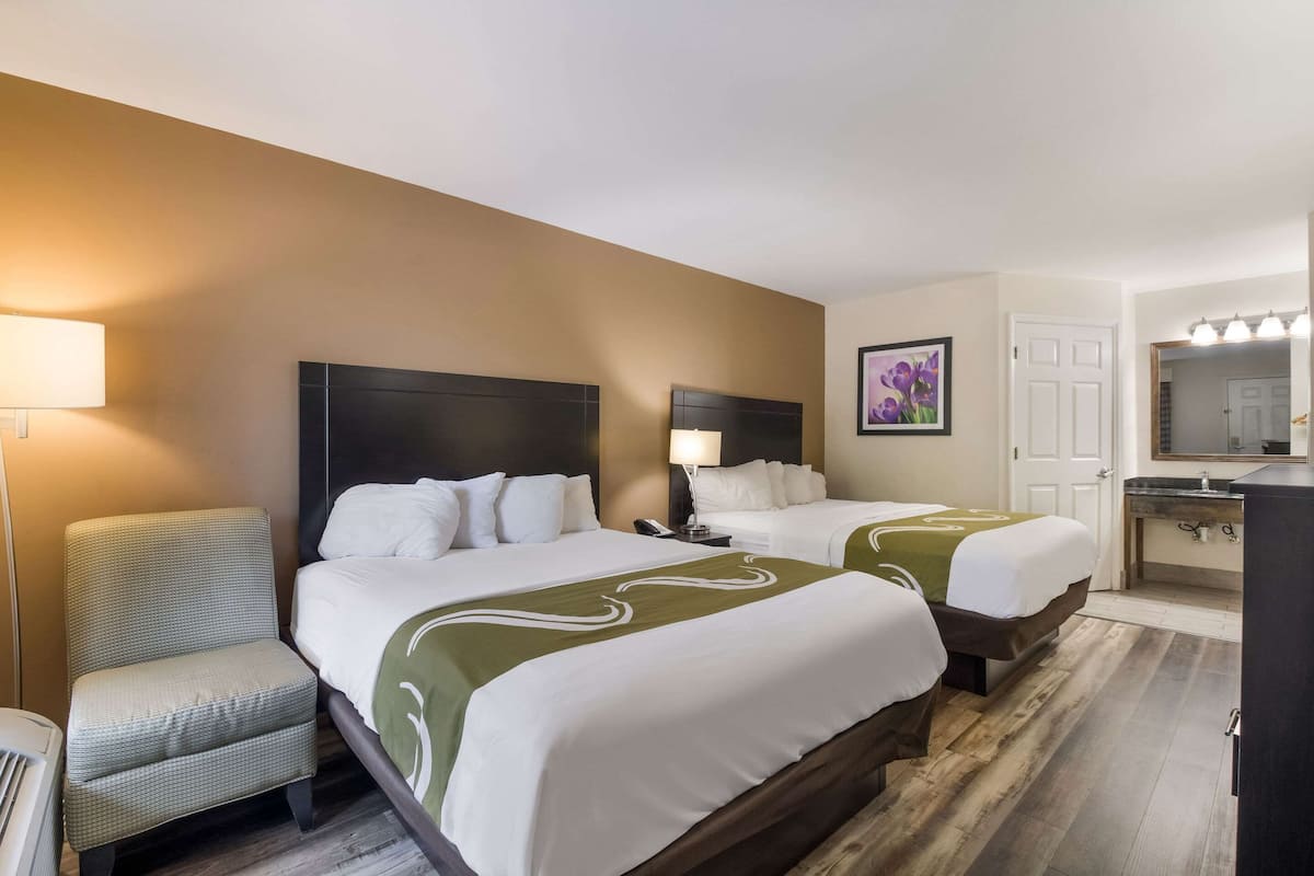 A hotel room with two beds, a chair, a desk, a mirror, and a bathroom.