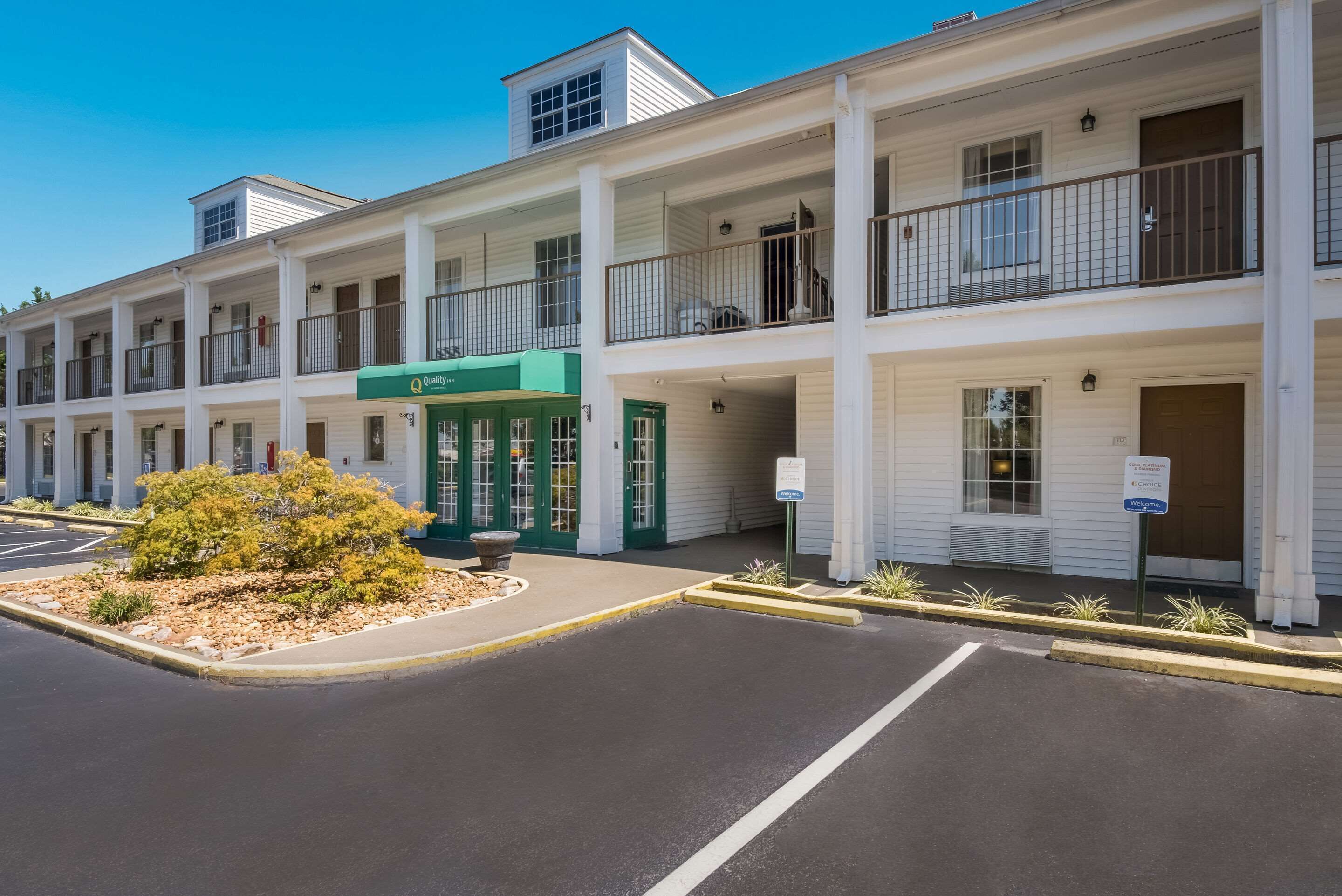 Un motel à plusieurs étages, avec une entrée verdoyante, un parking extérieur et un petit espace paysager agrémenté d’un arbuste.
