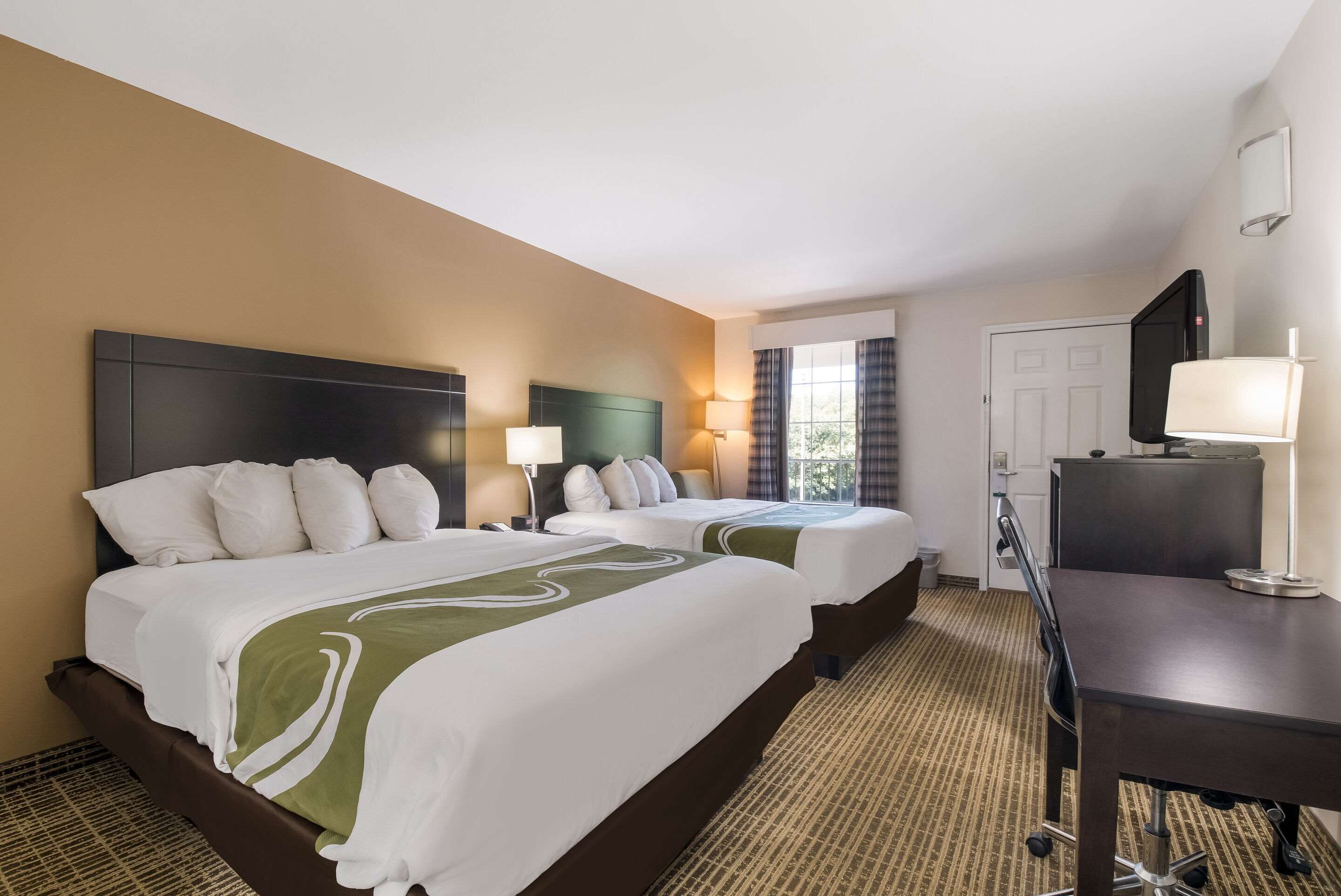 A hotel room with two beds, a desk, a television, and a window with curtains.
