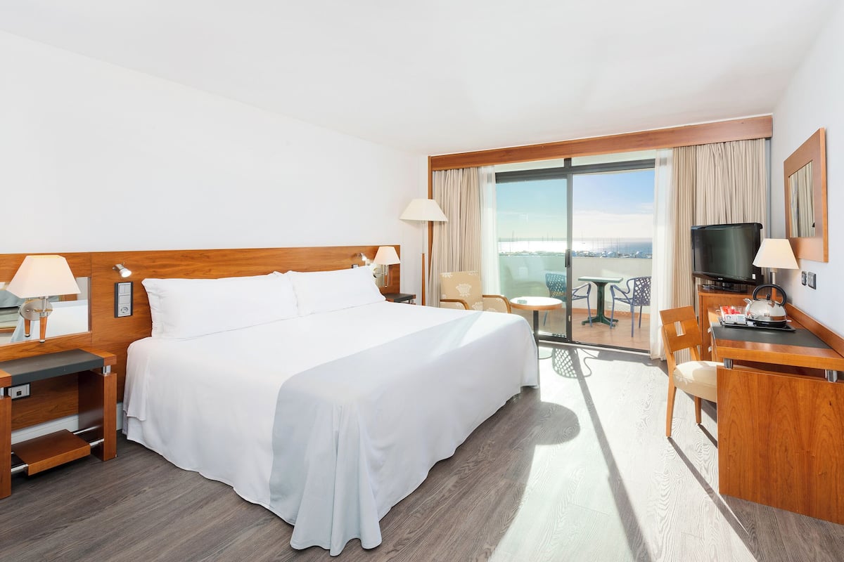 Ein Hotelzimmer mit einem großen Bett, einem Schreibtisch mit Fernseher, einem Sessel und einem Balkon mit Blick.