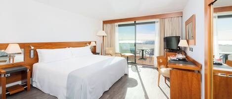 Habitación de hotel con una cama grande, un escritorio con televisor, una silla y un balcón con vistas.