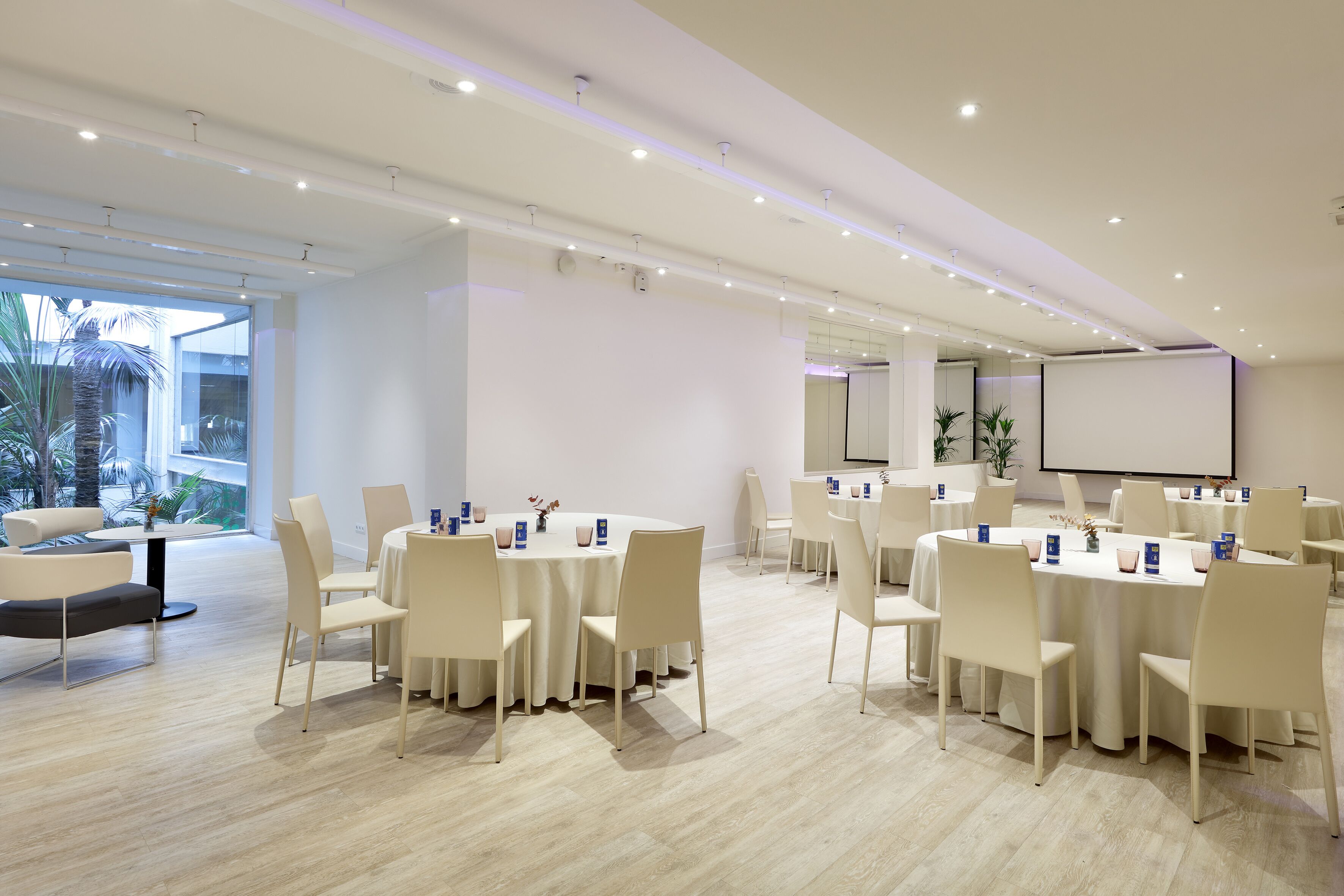 Una sala de conferencias moderna con mesas y sillas redondas, una pantalla grande y un balcón con plantas.