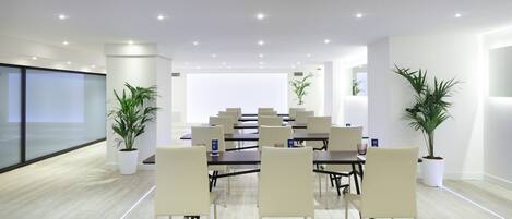 Un salón de conferencias moderno con sillas y mesas blancas, y plantas en macetas.