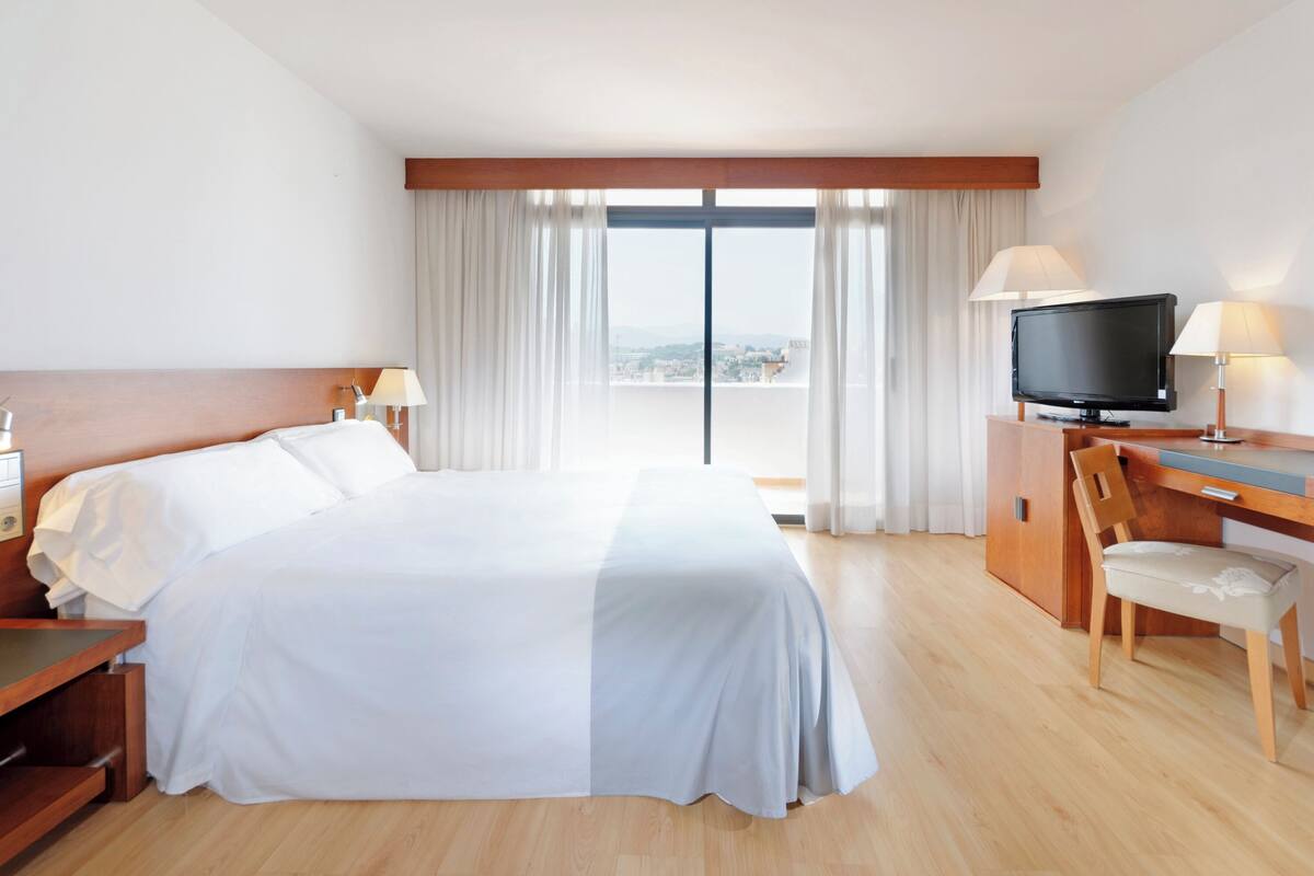 A hotel room with a large bed, a desk with a television, and a view of the city through the window.