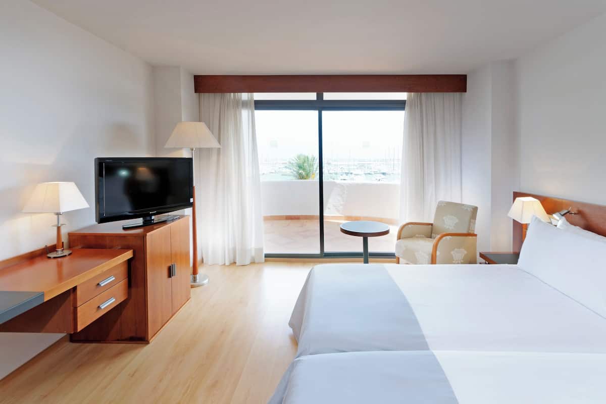 Ein Hotelzimmer mit einem Bett, einem Fernseher auf einer Holzkommode, einem Sessel, einem kleinen Tisch und Blick auf den Hafen durch große Fenster.