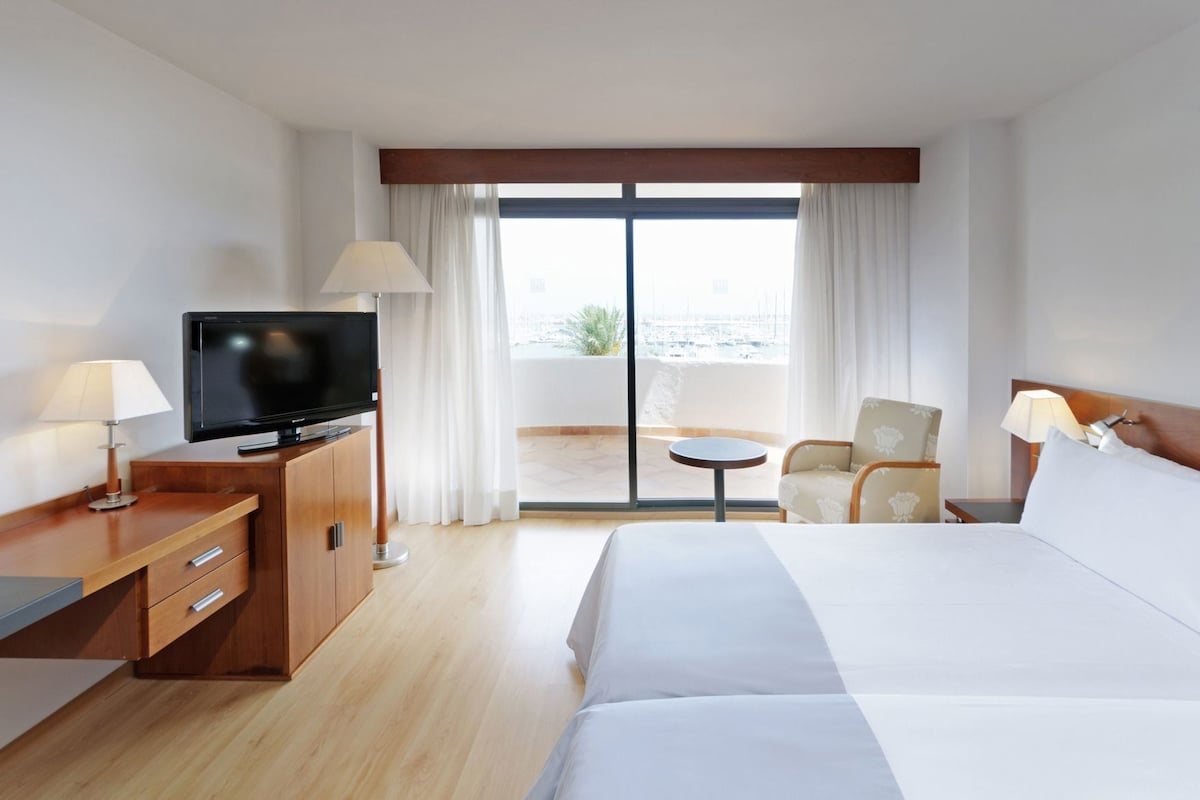A hotel room with a bed, a TV, a desk with drawers, a chair, a small table, and a balcony with curtains.