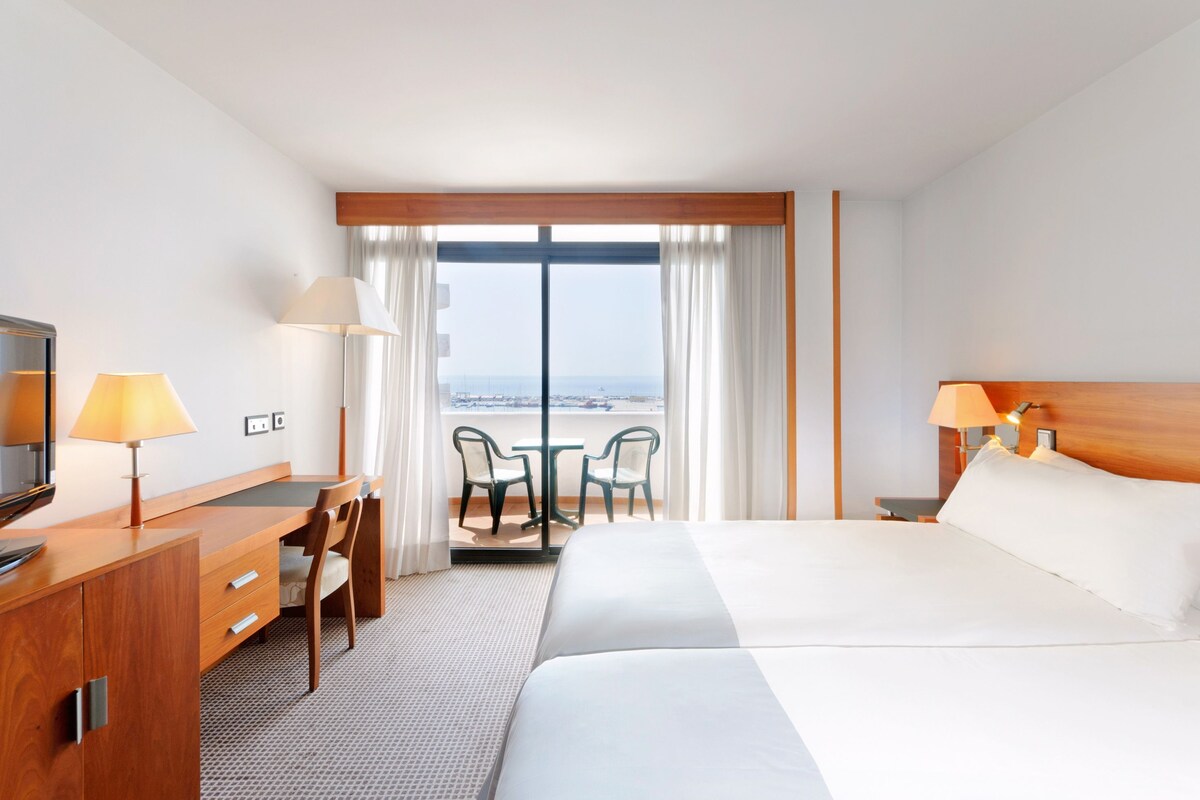 A hotel room with a bed, desk, chairs, and a view of the sea.