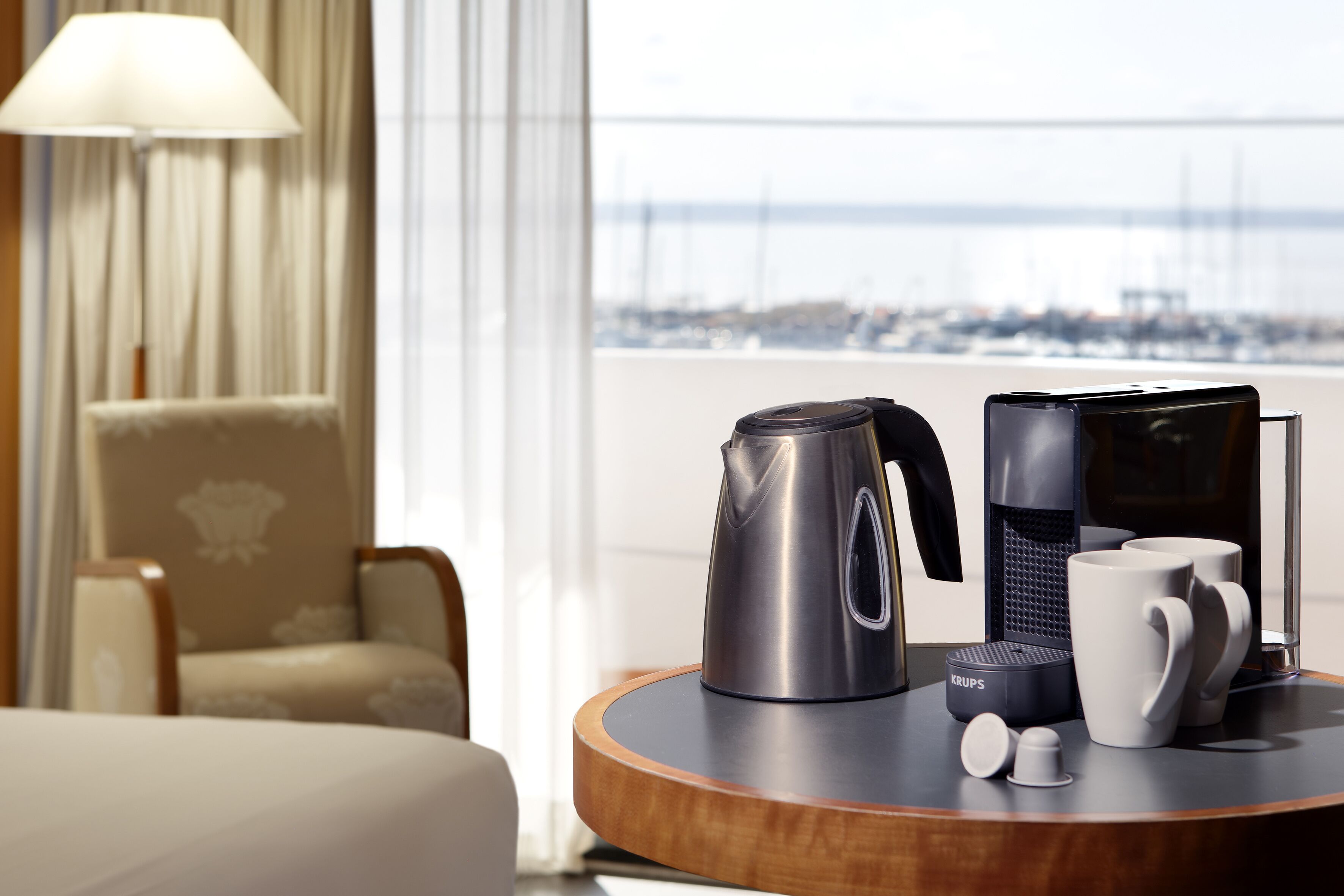 Habitación de hotel con vista al mar, cafetera, tetera y un molinillo de café Krups sobre una mesa de madera.