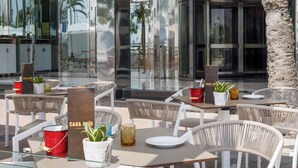 An outdoor seating area with wicker chairs and tables, a palm tree, and a glass wall reflecting the surroundings. - Hotel Palma Bellver Affiliated by Meliá (Palma de Mallorca)
