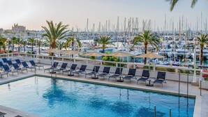 A swimming pool with lounge chairs, palm trees, and a marina in the background. - Hotel Palma Bellver Affiliated by Meliá (Palma de Mallorca)