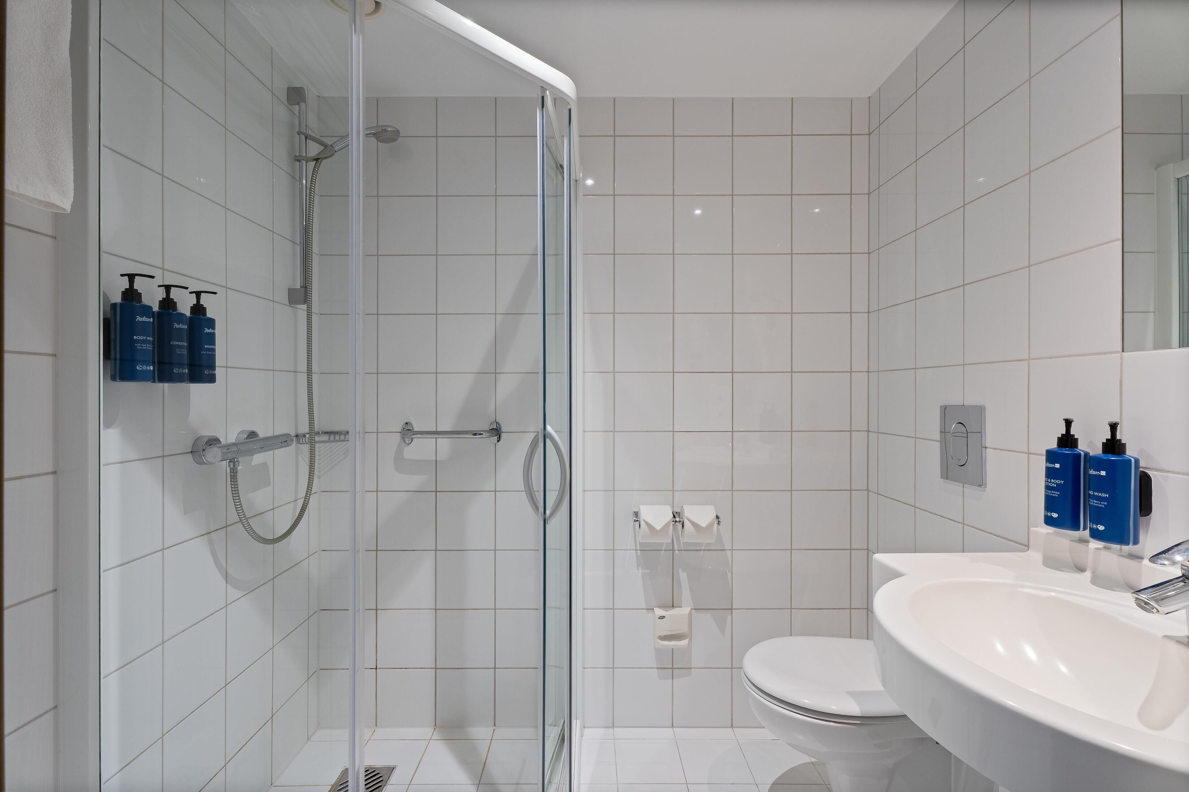 A modern bathroom with a glass-enclosed shower, white tiles, a white sink, and two bottles of shampoo on the wall.