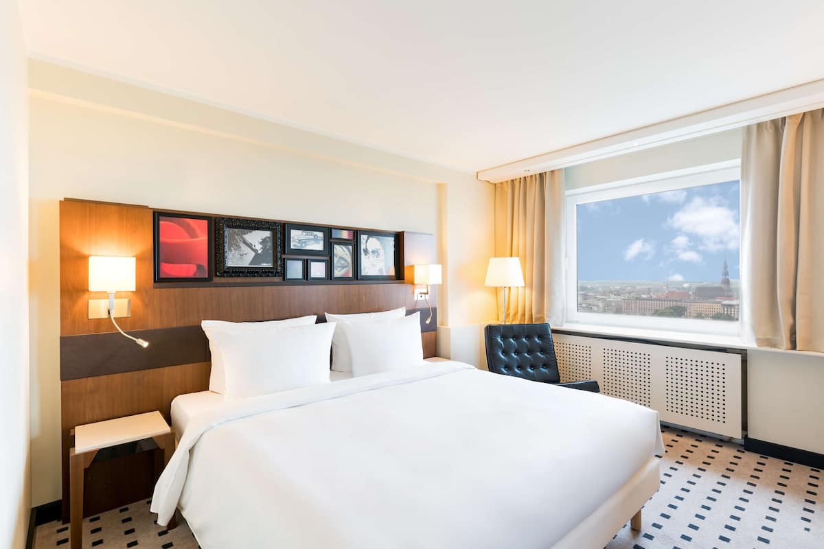 Habitación de hotel con una cama grande, dos lámparas de noche, cabecera con fotografías enmarcadas, ventana con vistas a la ciudad y un radiador.