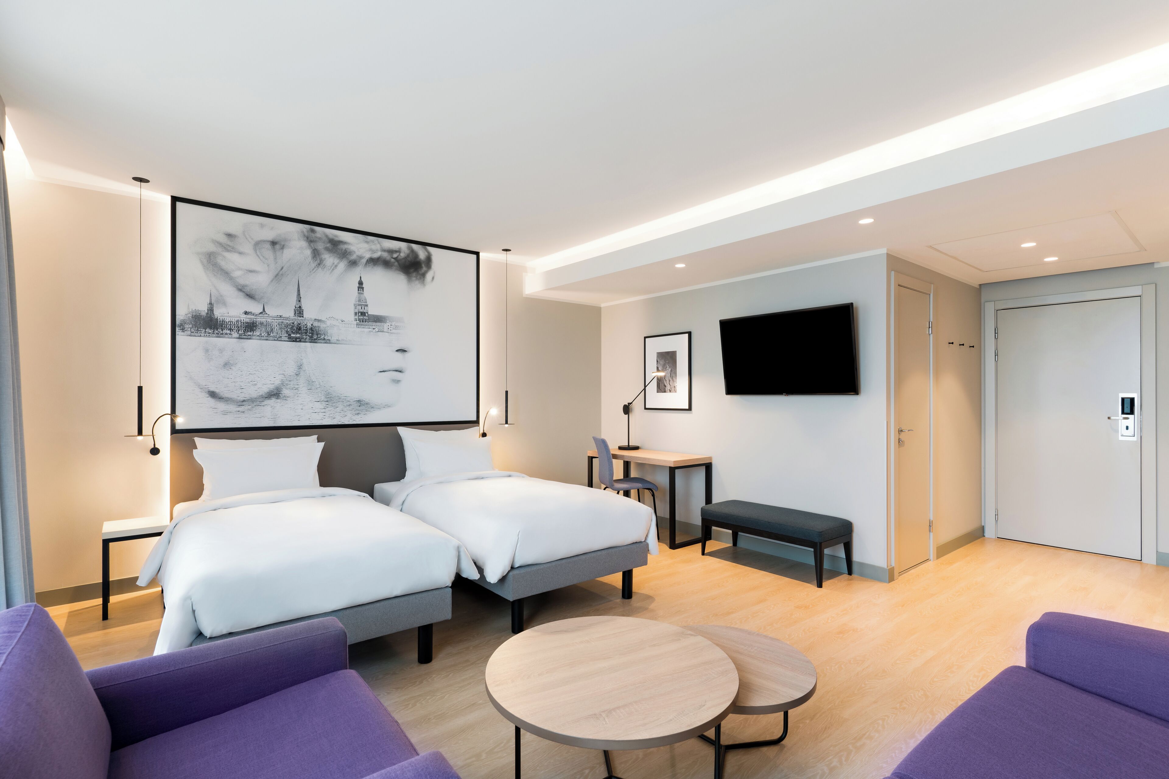 A modern hotel room with two beds, a desk, a chair, a round table, and a TV mounted on the wall.