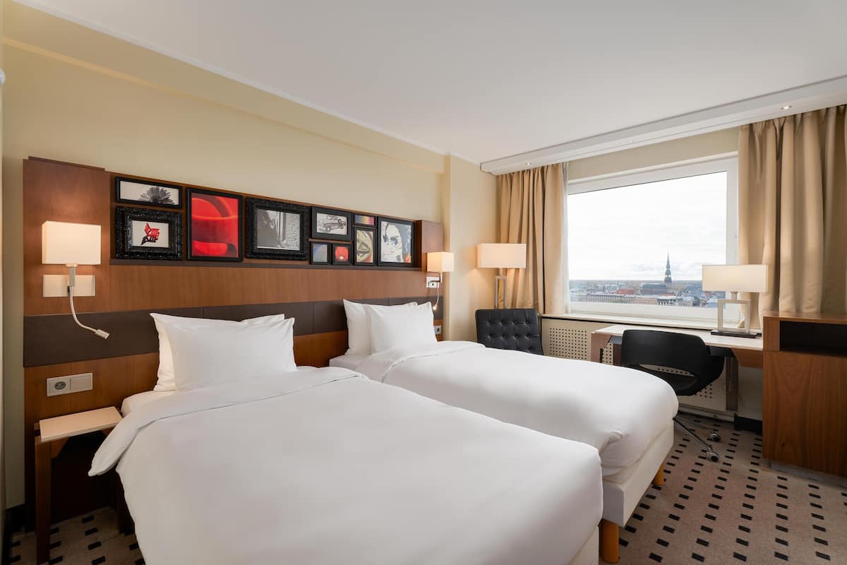 Habitación de hotel con dos camas, un escritorio, una silla y un amplio ventanal con vistas a un paisaje urbano.