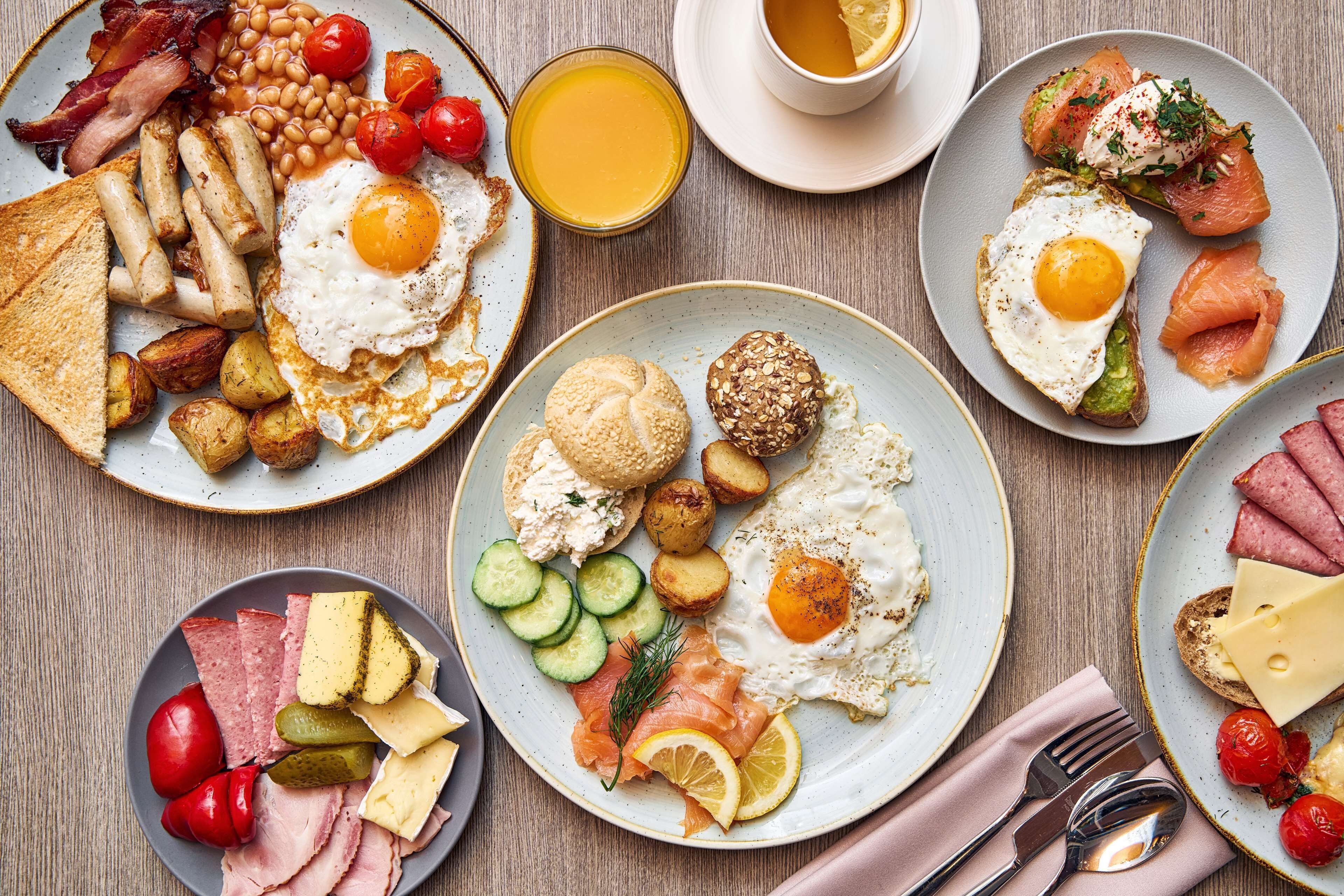 Una mesa de desayuno con diversos platos, incluyendo huevos, tocino, salchichas, tostadas, y una variedad de quesos y embutidos.