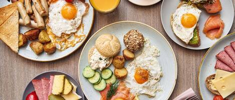 Una mesa de desayuno con diversos platos, incluyendo huevos, tocino, salchichas, tostadas, y una variedad de quesos y embutidos.