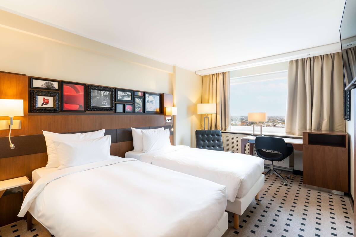 Habitación de hotel con dos camas, un escritorio, una silla, una ventana con cortinas y obras de arte colgadas en la pared.
