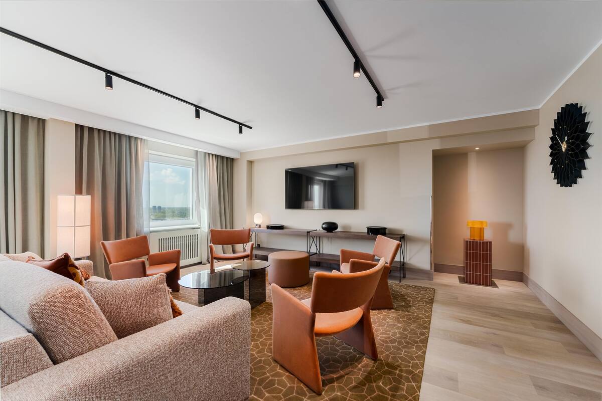 Una habitación de hotel moderna con sofá, sillones, una mesa de centro, televisor y un ventanal con cortinas.